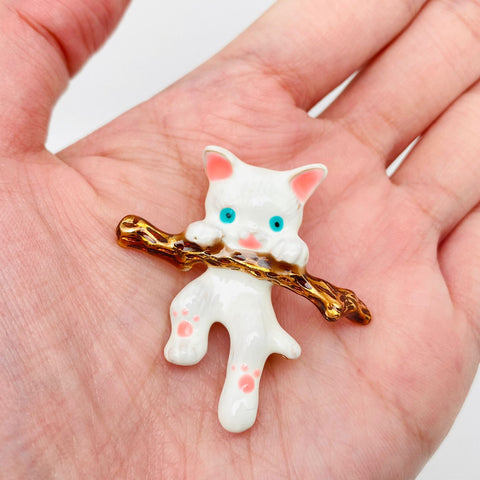 White, brown, blue, pink enameled gold metal brooch of a white kitten hanging from a branch. Shown up close in hand