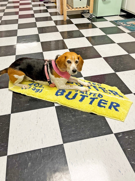 Rug shaped like a yellow stick of butter with blue lettering and a white outline. Shown on a checkered floor with a beagle 