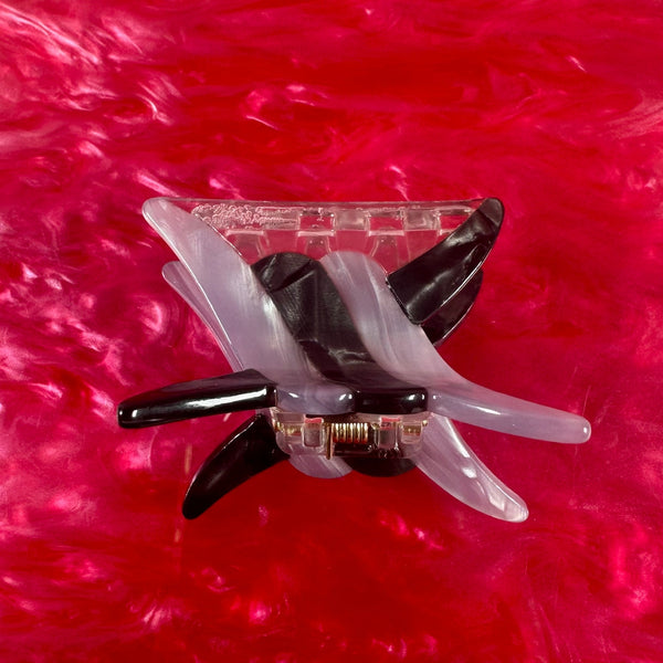 Black and grey marbled acetate hair clip in the shape of a link of barbed wire on a pink textured background. Shown from top