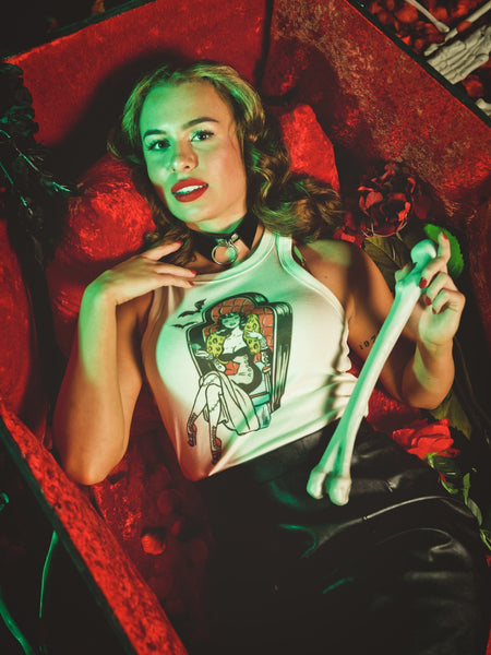 off-white ribbed tank top with round neck and printed illustration of pinup style woman with stitched legs wearing gown and sitting in coffin-style large chair. Printed in red, black, blue, and yellow. Shown on worn by model lying in coffin