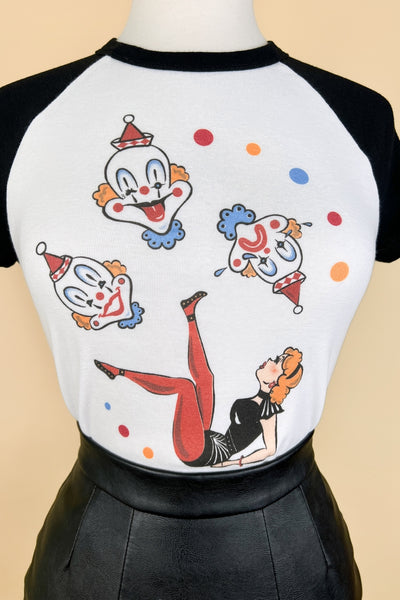 white crop t-shirt with black raglan sleeves & collar and printed illustration of woman in circus acrobat costume lying on her back with legs posed in the air surrounded by three large primary colored clown heads and primary colored dots. Shown on dress form in close up tucked into leather skirt