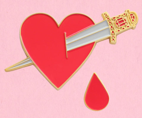 Set of two gold metal and red soft enamel pins in the shape of a large heart pierced by a dagger with an ornate gold handle and a small drop of blood. Shown on a light pink background