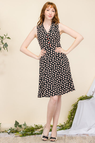 model wearing a sleeveless shirtwaist dress in a black background white and red daisy pattern. It has red plastic buttons running down the bodice and a v-neck shawl style collar. It has a slightly flared skirt ending at the knee. Shown from the front