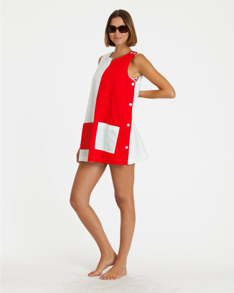 a model wearing a cotton a-line smock style mini dress in half red, half white design divided vertically down the middle of the garment. it has a round neckline with slash detail, button closure running down the left shoulder and down the side of the dress, and patch pockets in alternate colors at each hip. Shown from side
