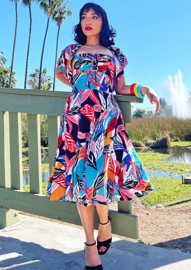 A model wearing a halter dress and bolero set in a bright blue, orange, pink, red, white, and black abstract tropical leaf pattern. It has a sweetheart neckline with exaggerated cuff details, cinched bodice, below the knee skirt, and adjustable halter tie neck. The bolero has short sleeves and a high mandarin style collar. Shown from the front