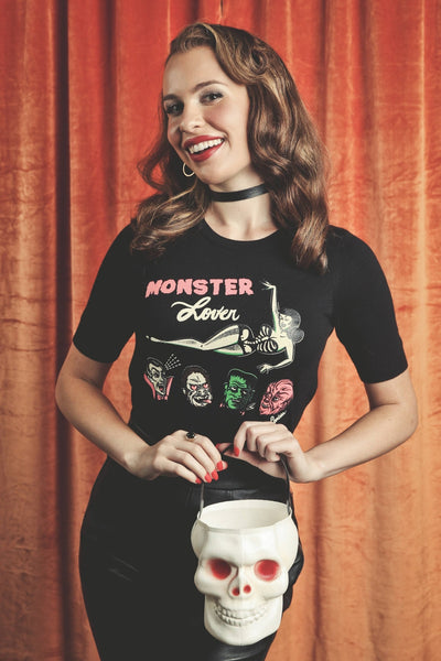 A woman wearing a black short sleeve pullover crew neck sweater with 'Monster Lover' text in pink and white and illustration of pinup style woman in black and white skeleton print swim suit and oversized heads of a vampire, wolfman, Frankenstein, and gorilla with hearts in their eyes. Shown holding a skull-shaped bucket against a textured orange curtain background.