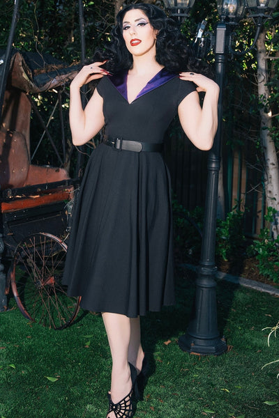 model wearing black bengaline swing dress with cap sleeves, v-neckline, shawl style collar in purple satin and oversized hood lined in matching satin. Dress has black faux leather belt and knee length full skirt. Shown from front