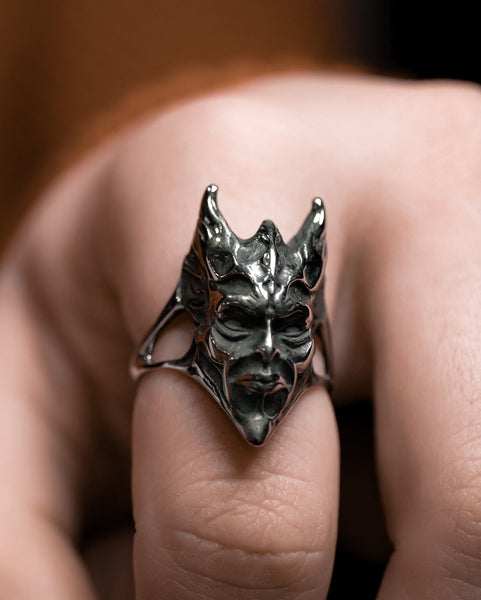 Model wearing stainless steel ring in the shape of a demon with pointed horns and face in angry expression with closed mouth. Seen in close up from the front 