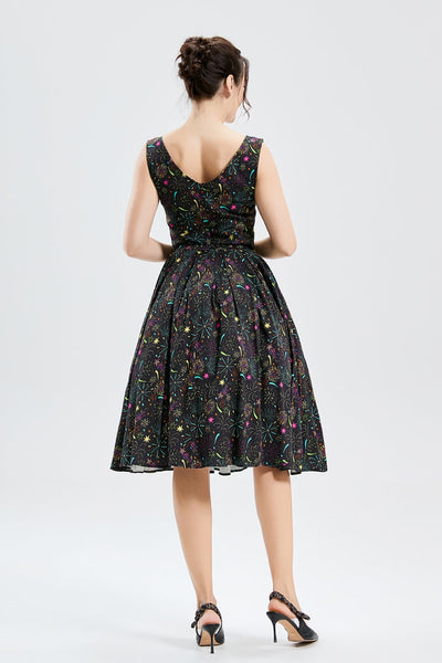 Model wearing a fit and flare dress made of soy silk in a black background pattern of exploding fireworks in bright red, pink, green, blue, orange, and purple. It has a high fitted neckline, princess seamed bodice, wide banded waist, and box pleated knee length skirt with pockets. Shown from the back