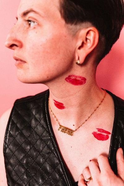 Gold covered stainless steel lip chain necklace with 'Freak' nameplate pendant shown worn by a model wearing a leather vest and red lipstick kisses on their tosrso