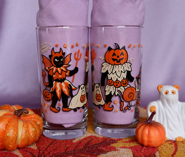 Drinking glasses screen printed with illustrations of cats wearing vintage inspired Halloween costumes. Two shown next to each other on a purple background
