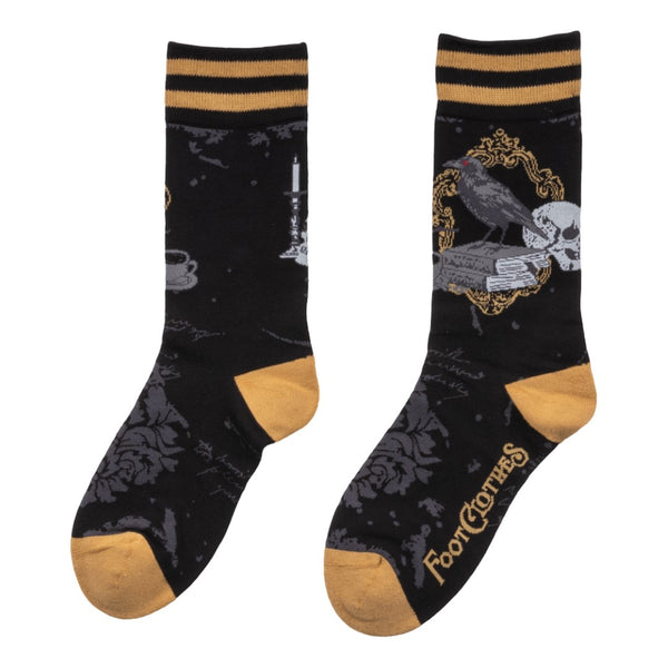 black unisex crew socks with a grey damask pattern, mustard and black striped cuffs, and mustard heels/toes. Ankle design is a raven perched on a stack of books alongside a skull, candlestick, and cup of tea. Shown flat