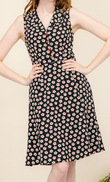 model wearing a sleeveless shirtwaist dress in a black background white and red daisy pattern. It has red plastic buttons running down the bodice and a v-neck shawl style collar. It has a slightly flared skirt ending at the knee. Shown from the front in close up of pattern