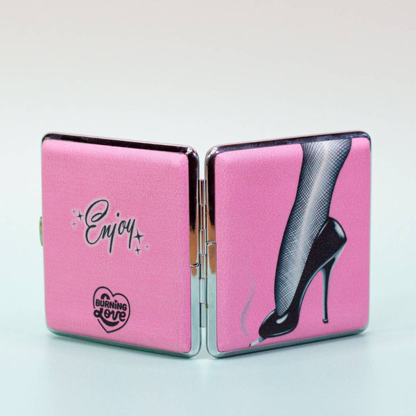 Square metal cigarette case with a printed pink front and black and white image of a high heeled shoe with fishnets stubbing out a cigarette. Shown open from the back with both sides visible. Back of the case has message “Enjoy!” with A Shop of Things logo