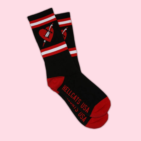 black unisex crew socks with red toes, heels, and white and red striped cuffs with red heart pieced by dagger on ankle. "HELLCATS USA" on bottom of sock