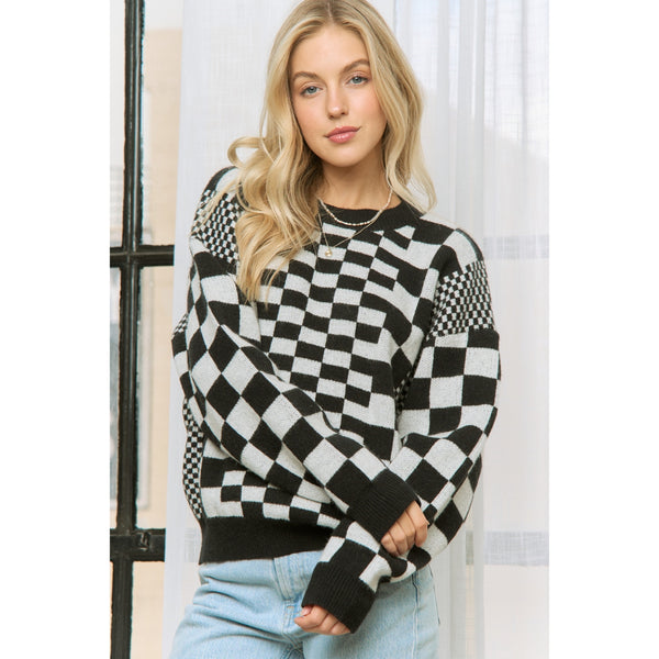 Woman wearing a black and white checkered sweater with various sizes of checker patterns and ribbed black collar, cuffs, and bottom hem. Shown from front