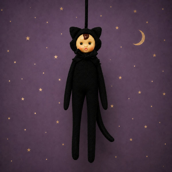 Poseable ornament with wire limbs and Velcro hands in the shape of a pixie with rosy cheeked, green eyed resin face with molded tuft of brown hair. She is wearing a black faux fur black cat costume with ears and tail. Black satin ribbon for hanging is at top of head. Shown on purple background with gold celestial pattern