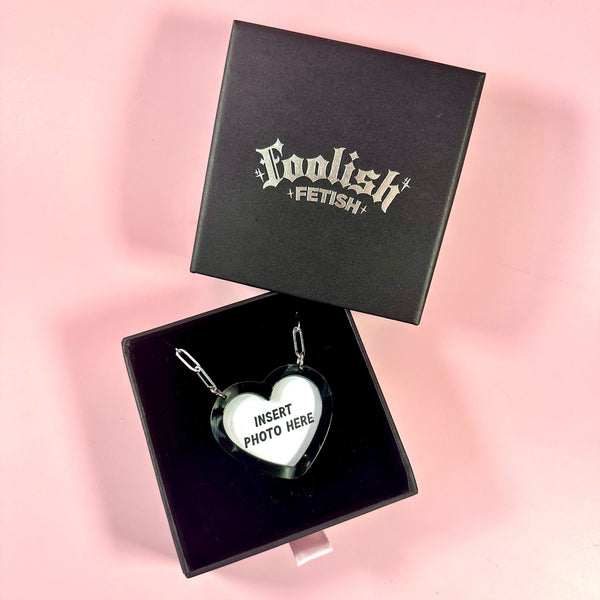A heart-shaped custom photo frame pendant in a black box with a black lid printed with the brand name 'Foolish Fetish' in silver gothic lettering, set against a solid pink background.