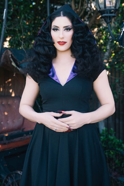 model wearing black bengaline swing dress with cap sleeves, v-neckline, shawl style collar in purple satin and oversized hood lined in matching satin. Dress has black faux leather belt and knee length full skirt. Shown from front with hood up