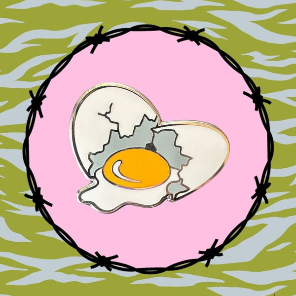 silver metal enamel pin of a white egg cracked in two pieces with a large yellow-yoked egg in between. Shown on barbed wire and blue/green zebra background