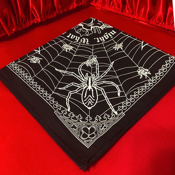 Square black cotton bandana with screen printed white image of mirrored bats and spiders inside a web with center image of vampire skull with quote “ Listen to them, the children of the night. What music they make!”. Shown folded to show only spider printed corner on red velvet background