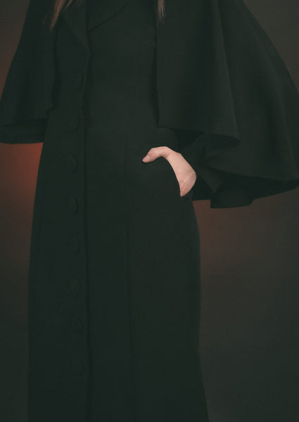 Model wearing black full length faux wool-blend coat with shawl collar and attached elbow-length cape. Showing close up detail of hand in side seam pocket