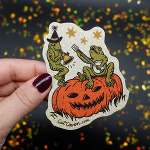 die cut vinyl sparkly sticker with illustration of two green frogs on top of a carved pumpkin playing a banjo and horn