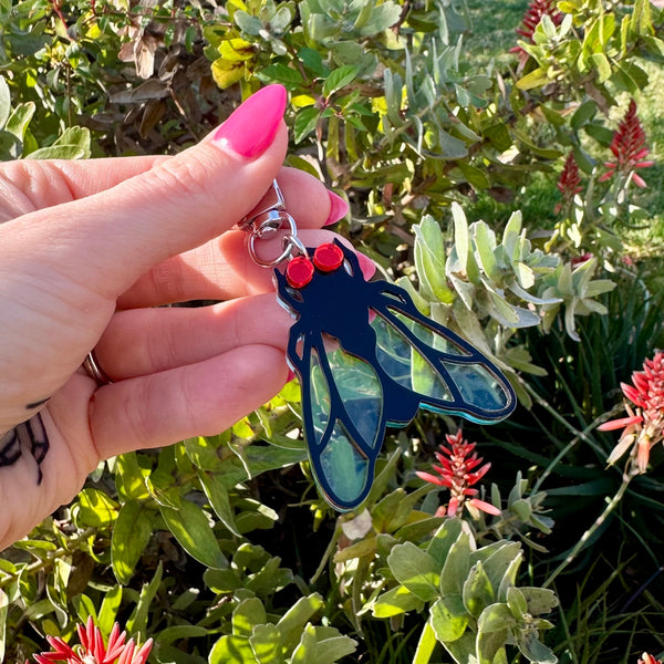 Laser-cut acrylic in the shape of a house fly with black body and transparent matte finish blue wings and red mirrored eyes. Attached to silver metal keychain via silver metal O-ring. Shown held in front of nature background for scale