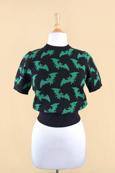 short sleeve acrylic knit sweater with black background and all-over tessellated bright green flying bat print. The top has a crew neck and ribbed hem. Shown from front