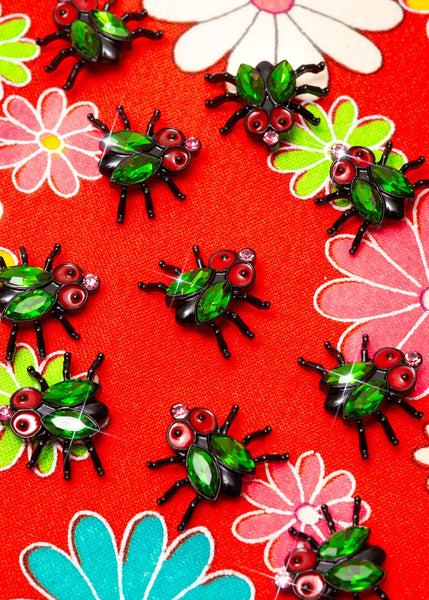 a group small shiny black enameled house fly-shaped broochs with enameled red eyes, almond-shaped bright green jewels for wings, an a small light pink jewel for a nose. Shown on red floral background