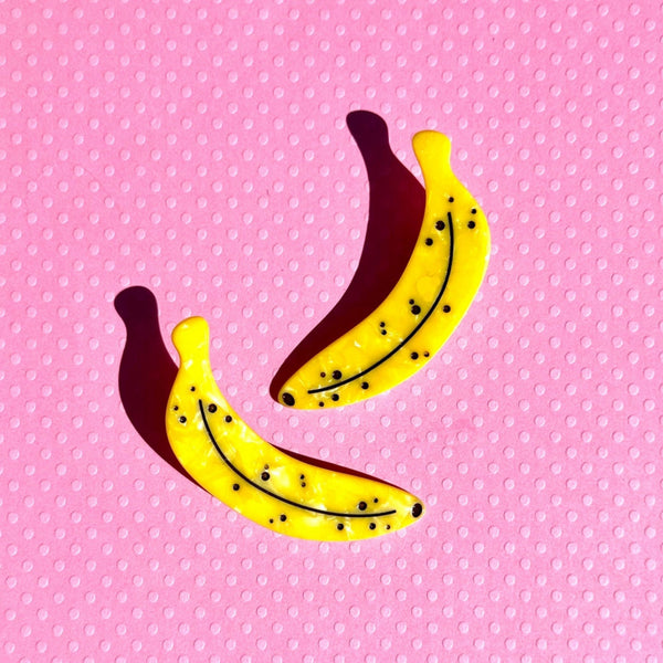 pair of acetate hair clips in the shape of bright yellow marbled ripe bananas with black spots. Shown on pink background