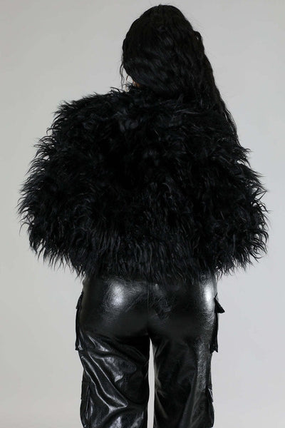 A model wearing a voluminous black shaggy faux-fur cropped open front jacket over a black top and shiny black faux-leather cargo pants. Shown from back