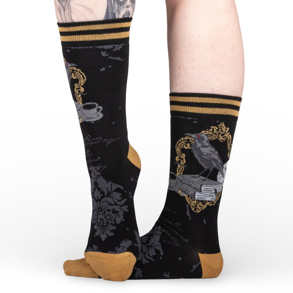 black unisex crew socks with a grey damask pattern, mustard and black striped cuffs, and mustard heels/toes. Ankle design is a raven perched on a stack of books alongside a skull, candlestick, and cup of tea. Shown worn from the side