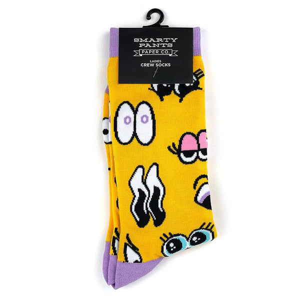 Crew socks with sets of cartoon eyes on a golden yellow background with lavender cuffs, heels, and toes. Shown flat in packaging from front