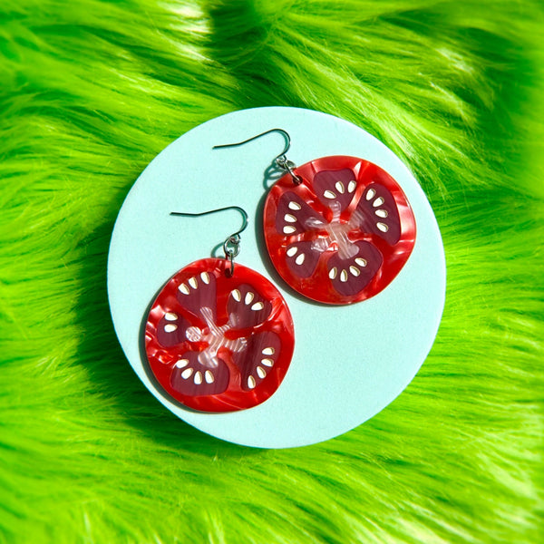 pair of marbled acrylic dangle earrings in the shape of slices of red tomato with seeds. Shown flat on green faux fur background