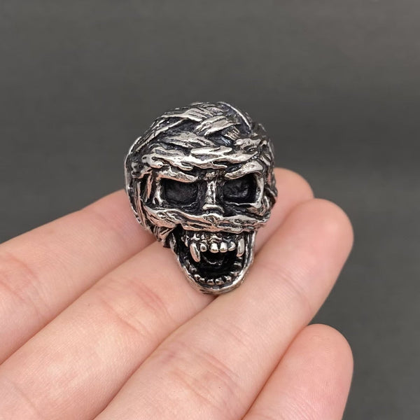 A silver stainless steel ring shaped like a mummy skull with snarling mouth, textured bandages and dark recessed details, shown resting on a person's fingers against a dark gray background. Shown from front