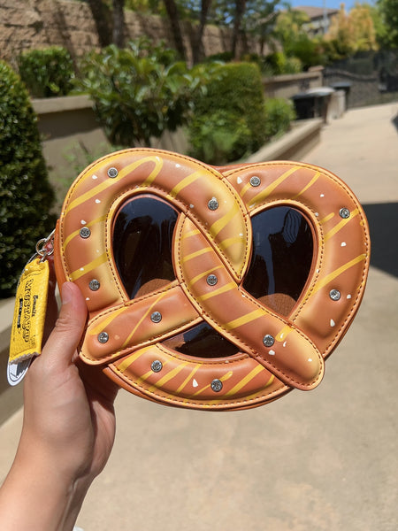 A crossbody purse in the shape of a large pretzel with mustard packet-shaped zipper pull, large rhinestones as grains of salt, and brown vinyl window. Shown from front held by hand in outdoor background