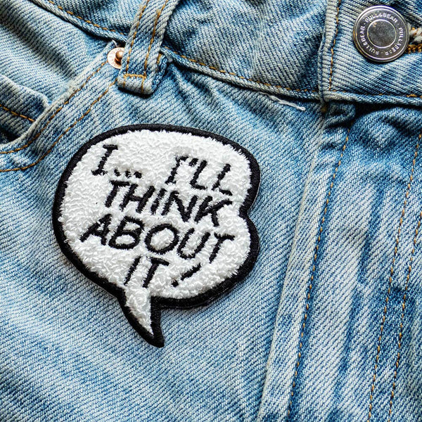 Embroidered patch with text 'I... I'll think about it!' in a speech bubble shape on white chenille background with black border. Shown on denim jeans