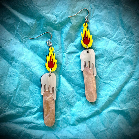 Laser-cut acrylic dangle earrings in the shape of a pair of burning melting candles. With bright yellow and red flames, pearl-finish white acrylic for the dripping wax, and glittery rose colored acrylic for the candle. Shown on a crumpled light blue paper background 