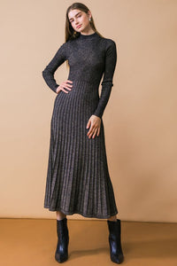 A model wearing a black and champagne gold lurex knit dress with high neckline, long sleeves, fitted waist, and below the knee pleated skirt. Shown from front