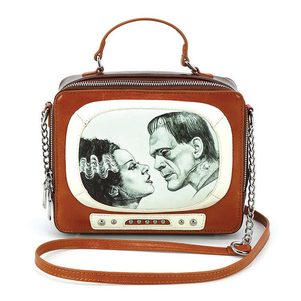 A rectangular novelty purse in the shape of a vintage television set in light brown and cream faux leather with silver details. Has a “screen” with a black and white glow-in-the-dark image of Frankenstein and his Bride and faux knob controls. Has a brown handle and detachable matching cross body strap. Shown from the front