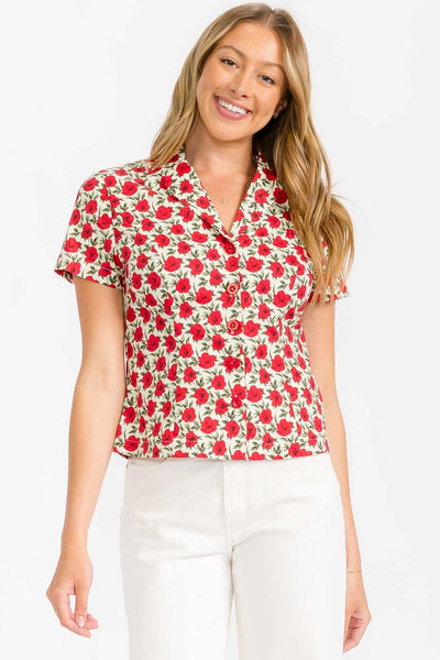 model wearing cotton short sleeve blouse with warm off-white background pattern of red and black flowers with green leaves. Has a notched collar and matching red plastic buttons. Shown from front