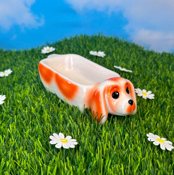 ceramic ashtray in the shape of a beige dachshund dog with orange details and black eyes & nose. The back of the dog is indented to form the tray area. Shown from side on fake green grass background