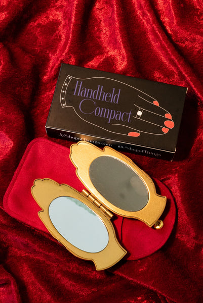 bright slightly burnished finish gold hand-shaped compact mirror with red enameled manicure and clear jeweled ring and bracelet detail. shown open on red velvet background alongside faux suede carrying case and gift box