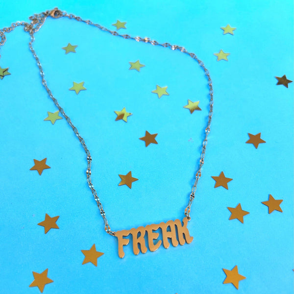 Gold covered stainless steel lip chain necklace with 'Freak' nameplate pendant on a blue background with gold stars
