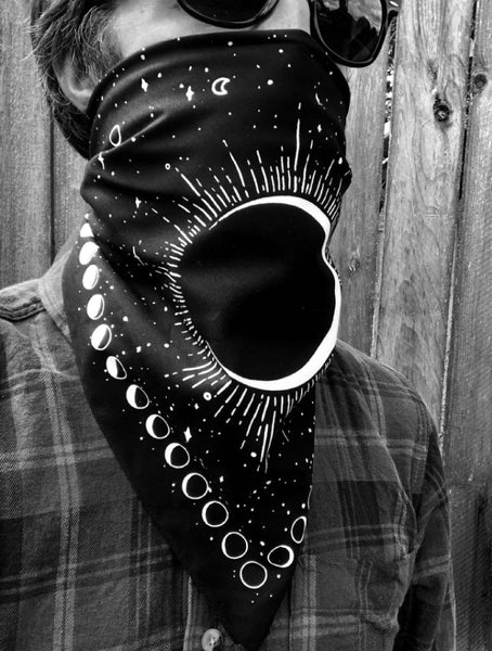 black cotton bandana with white screen printed design of large full moon opposite large crescent moon on starry background with border of moon phases. Shown worn covering mouth of person with sunglasses