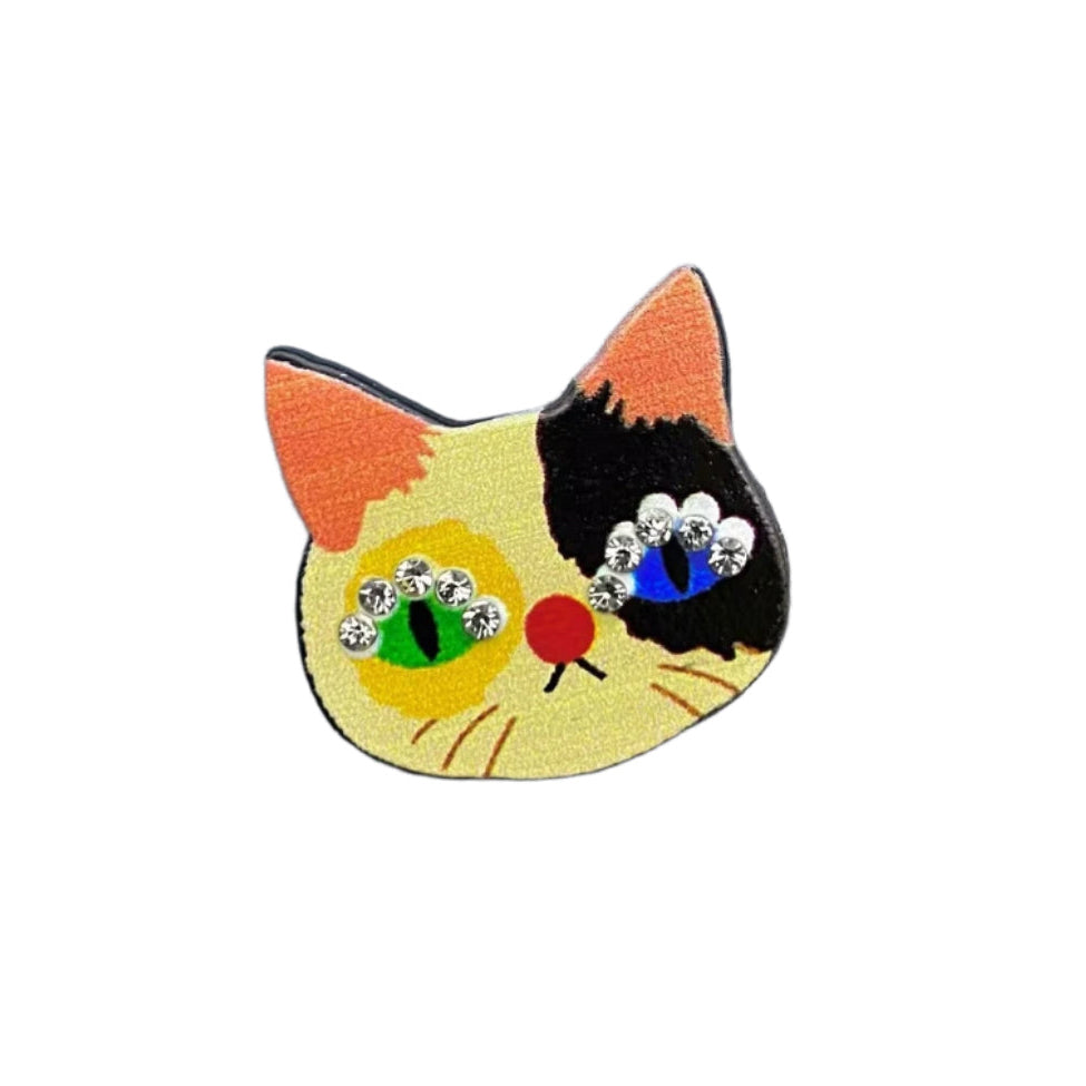 Small calico cat head brooch in black metal with printed details of black/cream/orange fur, pink ears, one green and and one blue eye, and 4 small silver rhinestones above the eyes
