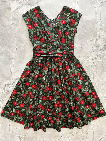 Mid length cotton dress with surplice neckline, cap sleeves, wide cummerbund style waistband, and full skirt. In a red and green holly, poinsettia, and mistletoe pattern on a black background. Shown flat