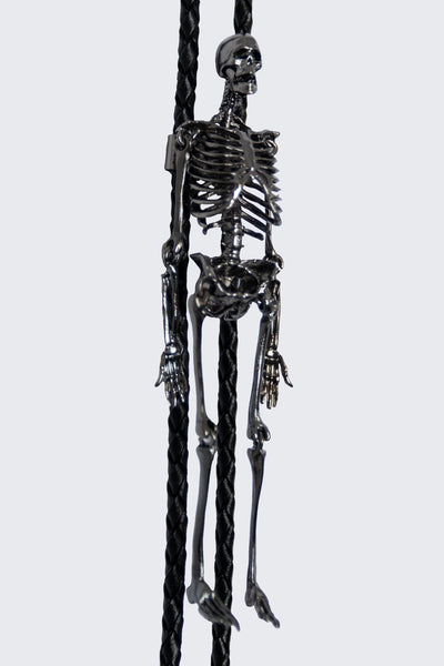 Black braided faux leather bolo tie with black metal tips and slide made of detailed gunmetal finish metal skeleton with articulated joints and head. Shown from the front in close up of skeleton
