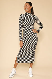 A model wearing a black and creamy white checkerboard pattern ribbed knit dress with long sleeves and a mock neck. Ends below the knee and has doubler slide slits. Shown from front